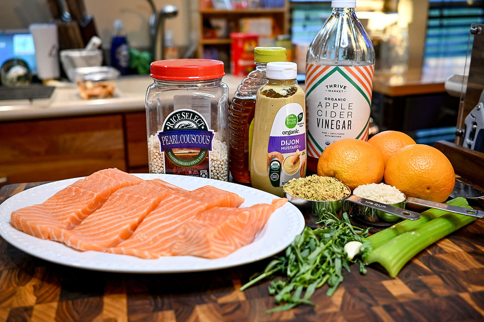 Salmon fillets, couscous jar, mustard, honey, vinegar, oranges, celery, and herbs on a kitchen counter with a wooden cutting board.