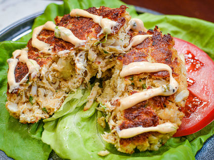 Crab cakes drizzled with sauce sit on fresh lettuce with a tomato slice. The golden crust contrasts the green and red background.