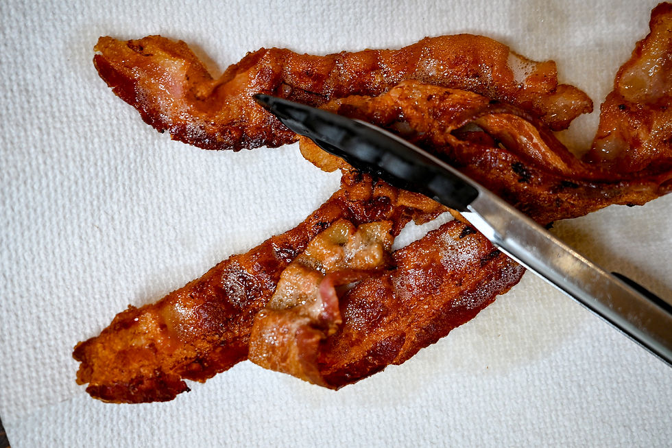 Crispy bacon slices on a white paper towel with black tongs resting on top. The setting is simple, focusing on the texture and color.