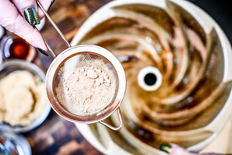 A hand with green nails sifts cocoa powder over a spiral-patterned bowl on a wooden table. Blurred ingredients appear in the background.
