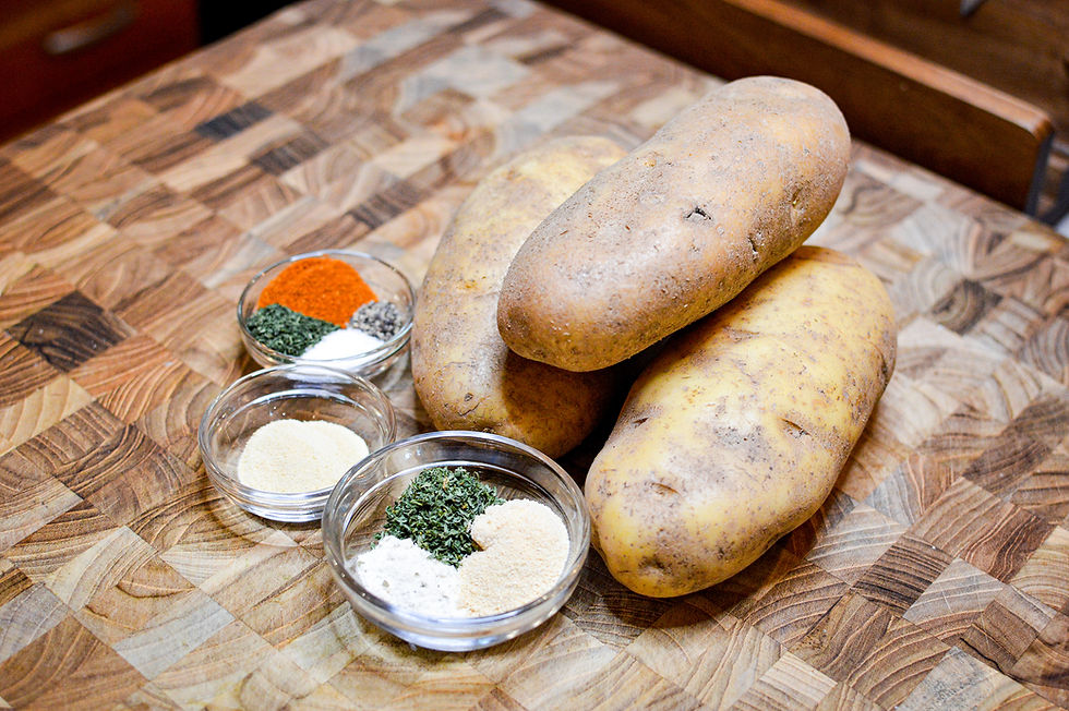 Potatoes on a wooden cutting board with small glass bowls of colorful spices, including paprika, parsley, and garlic powder. Warm kitchen setting.
