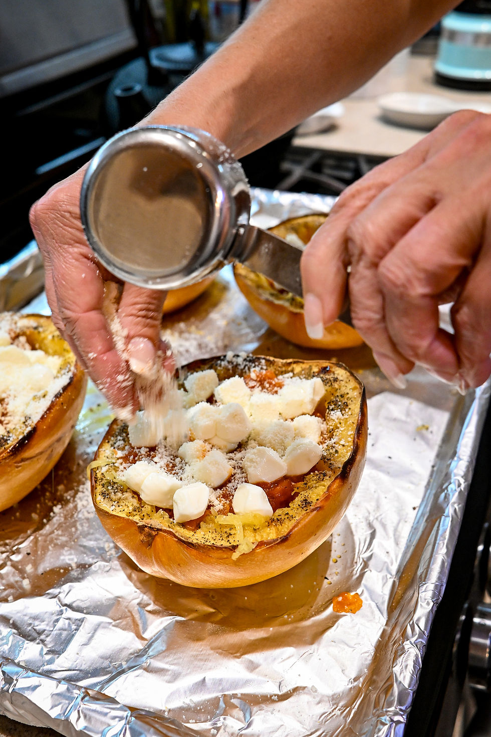Hands sprinkle grated cheese over stuffed roasted squash halves on a foil-lined tray in a kitchen, close-up food prep scene