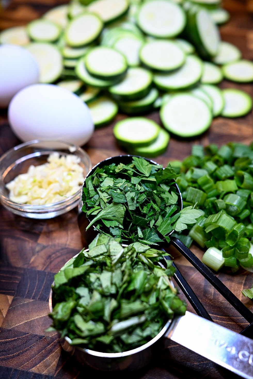 Chopped greens, sliced zucchini, raw eggs, and garlic on a wooden board. Fresh and vibrant cooking ingredients arranged in a kitchen setting.