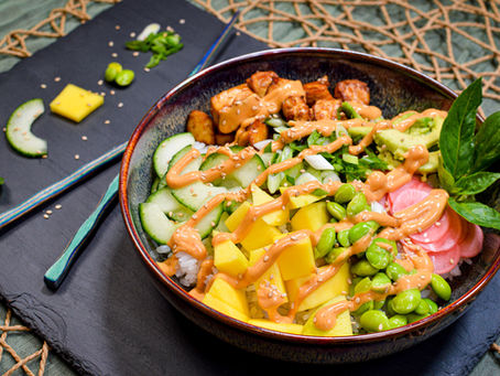 Colorful poke bowl with avocado, tofu, mango, edamame, cucumber, topped with sesame seeds and spicy orange sauce on a dark table.