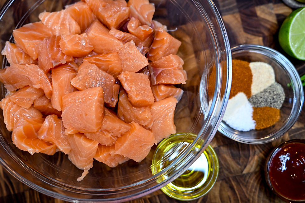 Chunks of raw salmon in a glass bowl on a wooden surface. Nearby, spices, lime, oil, and sauce are arranged in small bowls, ready for cooking.