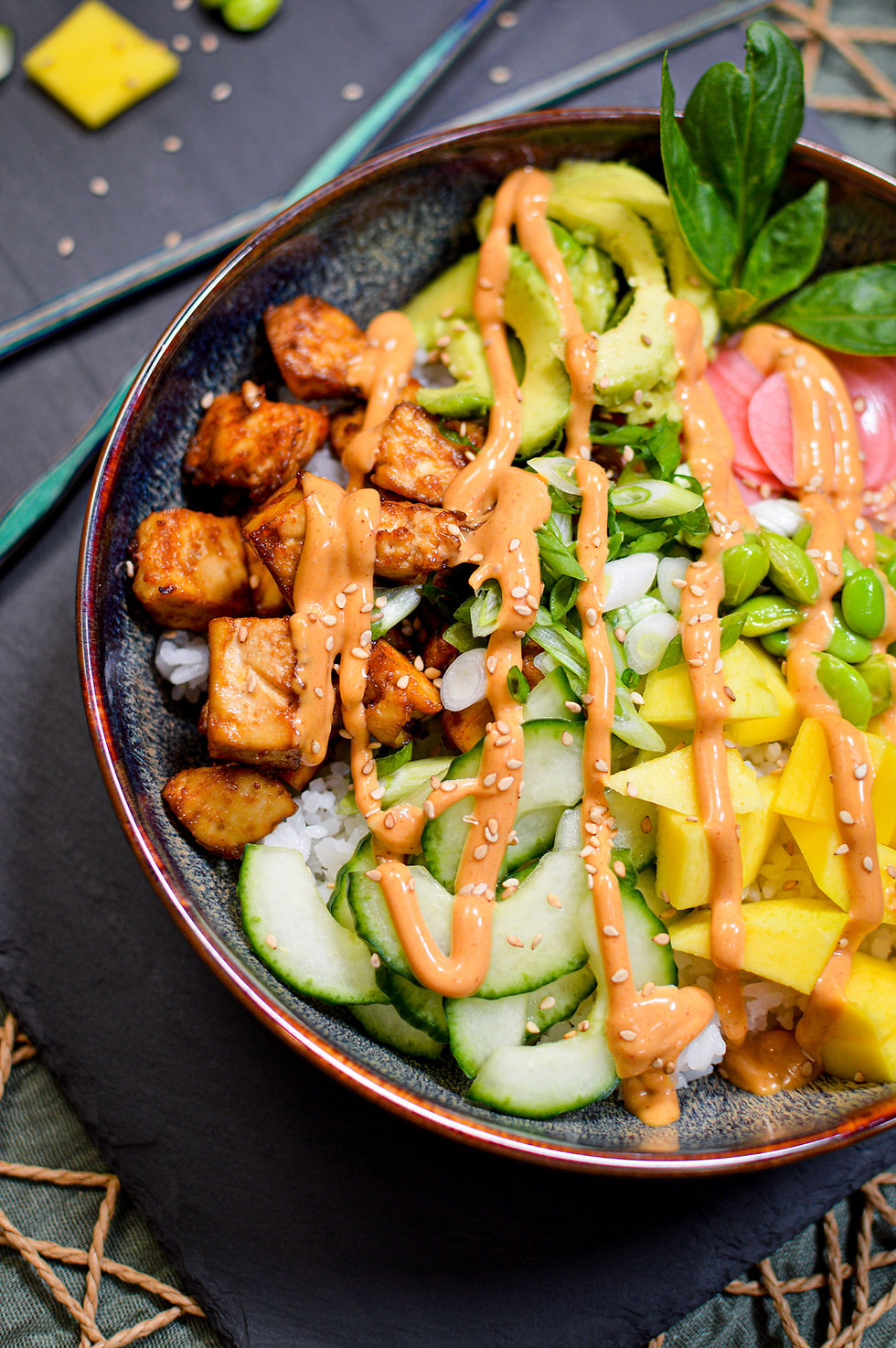Colorful poke bowl with avocado, tofu, mango, edamame, cucumber, topped with sesame seeds and spicy orange sauce on a dark table.