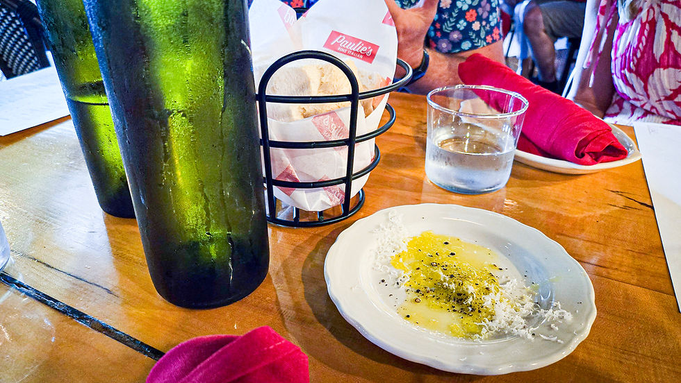 Green bottles, bread basket labeled "Paulie's," a plate with oil and spices, a glass of water, and red napkins on a wooden table.
