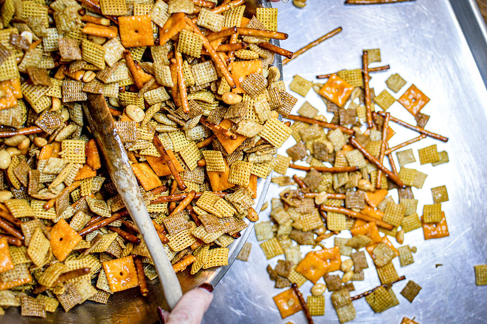 Snack mix with pretzels, cereal, cheese crackers, and nuts on a metal tray. A hand stirs the mix with a wooden spoon.