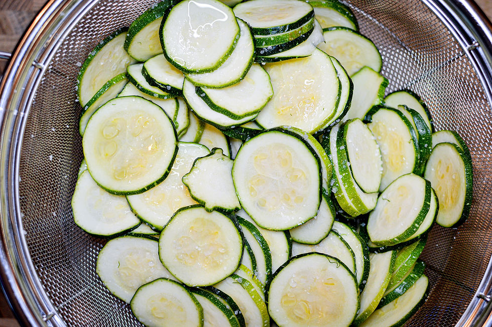 Sliced zucchinis with green edges in a metal colander, glistening with water droplets. The setting has a fresh and vibrant feel.