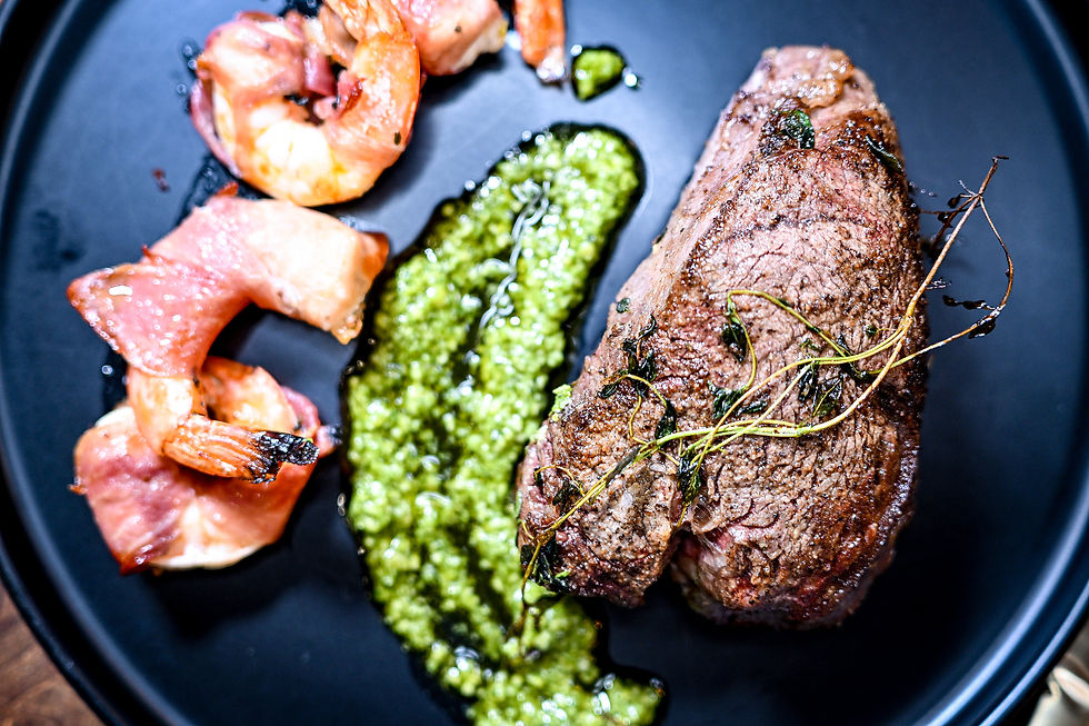 Steak with herbs, bacon-wrapped shrimp, and green pesto sauce on a black plate. Close-up shot, highlighting rich textures and colors.