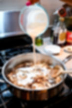 Milk being poured into a skillet with sliced mushrooms on a stove. Honey bottle and spices on the counter in the background. Cozy kitchen scene.