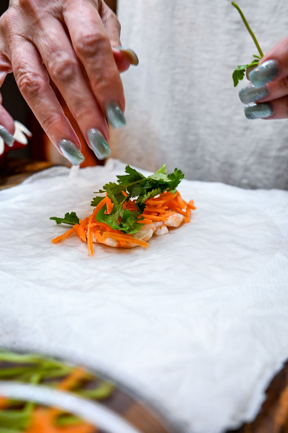 Hands with blue nails garnish a shrimp dish with carrot shreds and cilantro on white parchment.