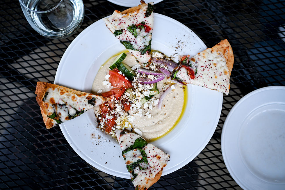 Pizza slices with cheese and basil surround a plate of hummus topped with tomatoes, onions, and feta on a black wire table.