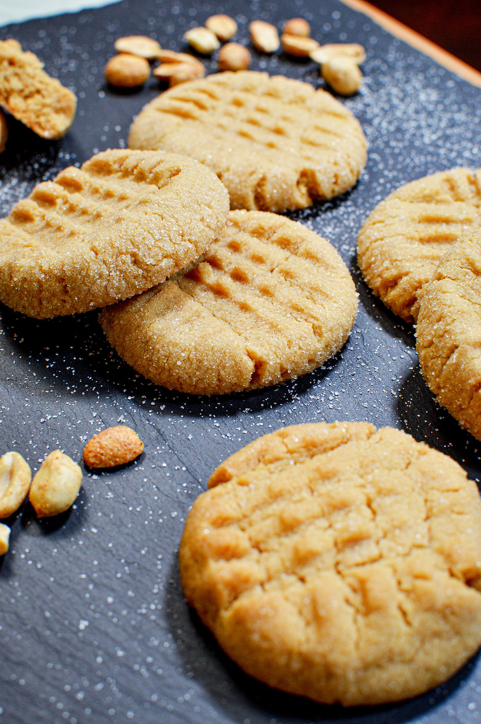 Peanut butter cookies on a dark slate, sprinkled with sugar, surrounded by shelled peanuts, creating a warm, inviting mood.