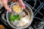 Hand with pink nails adds minced garlic to a saucepan of onions and green peppers on a stovetop.