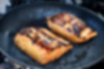 Two grilled salmon fillets with charred marks sizzle in a black frying pan, glistening with oil. The scene is in a kitchen setting.