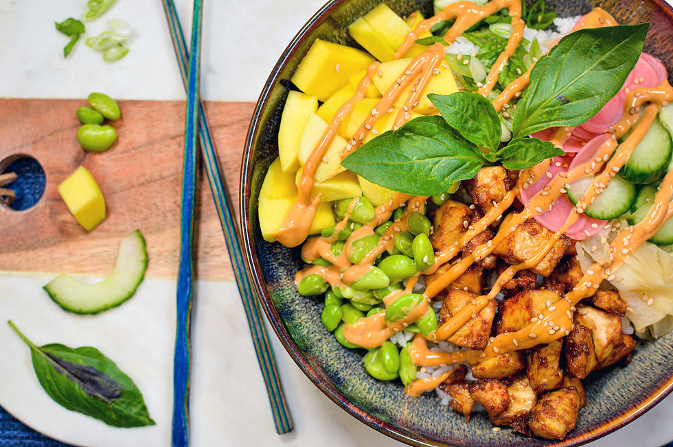 A colorful poke bowl with tofu, mango, edamame, cucumber, and greens, drizzled with sauce. Chopsticks rest on a wooden board.