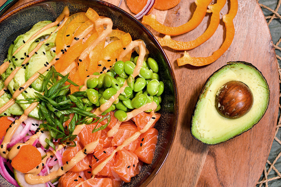 A colorful poke bowl with salmon, avocado, edamame, carrots, and sesame seeds on a wooden board. Half avocado on the side.