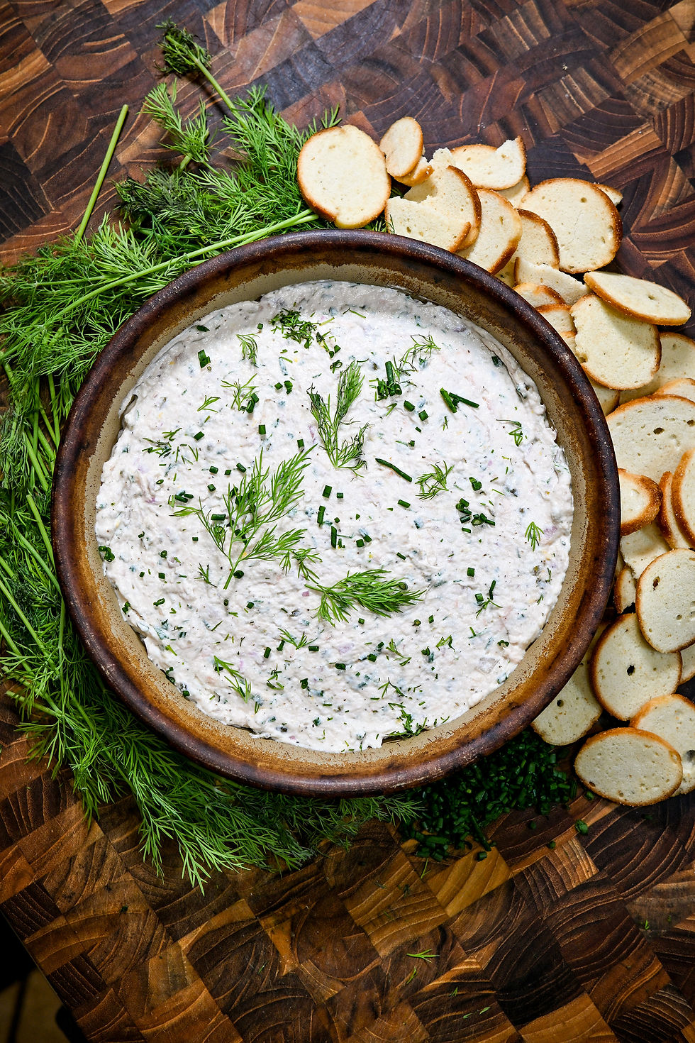 Creamy herb dip topped with dill and chives, surrounded by toasted bread slices on a dark wooden board.