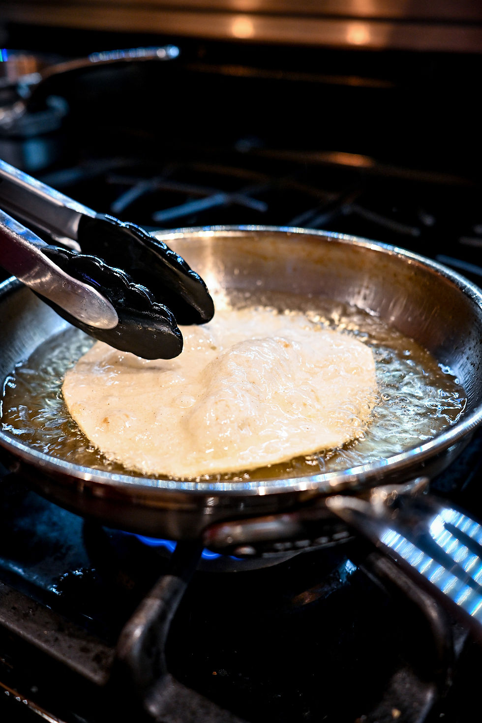 Tongs lift a tortilla frying in bubbling oil over a blue gas flame in a dark kitchen.