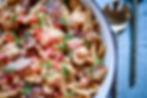 Close-up of creamy pasta with sausage, shrimp, tomatoes, and green onions in a white bowl. A golden spoon and fork rest beside it.