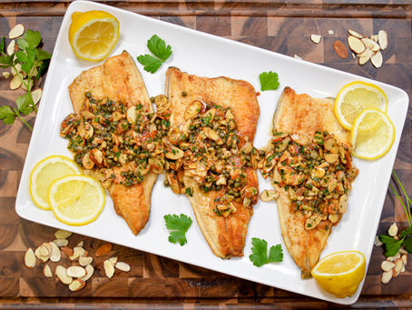 Grilled fish fillets topped with herbs and almonds on a white plate, garnished with lemon slices and parsley, set against a wooden surface.