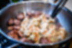 Shrimp and sliced sausage are being sautéed in a stainless steel pan with tongs lifting a piece, creating a sizzling, appetizing scene.