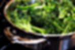 Fresh broccoli cooking in a shiny stainless steel pan on a stovetop. The vibrant green contrasts with the metallic surface.