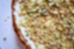 Close-up of a pie with a white frosting, topped with crushed pistachios. Edges are golden brown. Background is a plain white surface.
