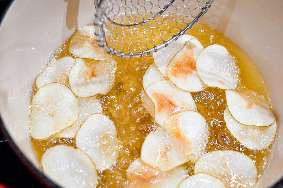 Potato slices frying in hot oil with a metal skimmer, in a pot. Golden oil, bubbles around slices. Crisping in progress.