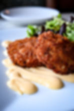 Crispy fish cakes on a white plate with creamy sauce, accompanied by a fresh green salad. Elegant dining setting, no text visible.