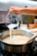 Hand pouring liquid from a Pyrex cup into a metal pan on a stove; wooden spoon stirs. Kitchen setting with jars in background.