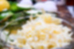 Close-up of grated cheese in a glass bowl, surrounded by sliced cucumbers, lemons, and eggs on a wooden surface. Bright and fresh setting.