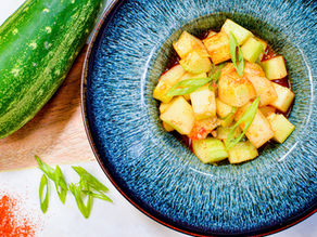 Diced cucumber salad in a blue bowl, garnished with green onions. A fresh cucumber and chili powder are on a wooden table background.