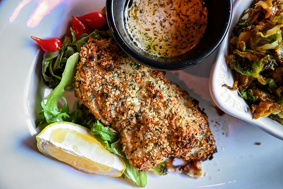 Herb-crusted fish with lemon on greens, surrounded by peppers and a bowl of sauce, with sautéed veggies on a white plate. Vibrant colors.