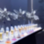 Perfume bottles on a lit table with white floral sculptures in the background. Wall text reads "INFINIMENT COTY PARIS" with additional phrases.