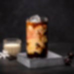 Iced coffee with swirling cream in a glass on a marble coaster, dark background, nearby white flowers, vanilla pod, and cherries.