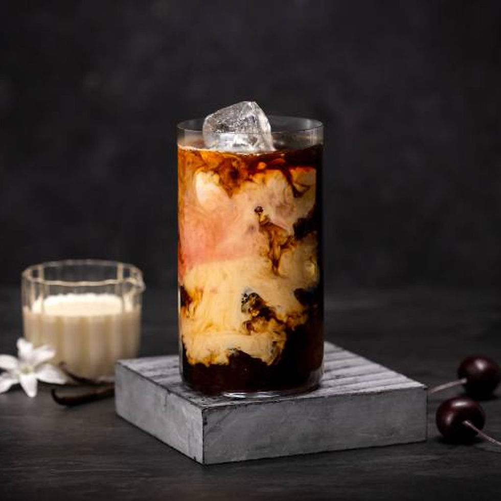 Iced coffee with swirling cream in a glass on a marble coaster, dark background, nearby white flowers, vanilla pod, and cherries.