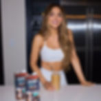 Smiling woman in white top holds iced coffee in a kitchen. Two cartons of Violife Sweet Cream are on the counter. Stainless steel fridge in background.