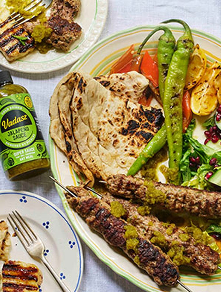 Grilled kebabs with green sauce on a platter with flatbread, grilled peppers, and salad. Bottle of Vadasz Jalapeno Sauce nearby.