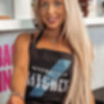 Smiling woman with long blonde hair wearing "My Protein Kitchen" apron. Bright kitchen background with visible product boxes and pink text.