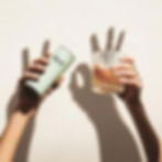 Hands holding a green "TRIP" can and a glass, against a beige wall. Shadows form an "OK" gesture. Bright, minimalist setting.