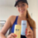 Smiling woman in a blue top and black cap holds two TRIP cans, one yellow and one blue, in a bright indoor setting.