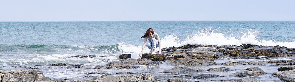 Woman collecting sea water on a rocky coastline with waves splashing around her.
