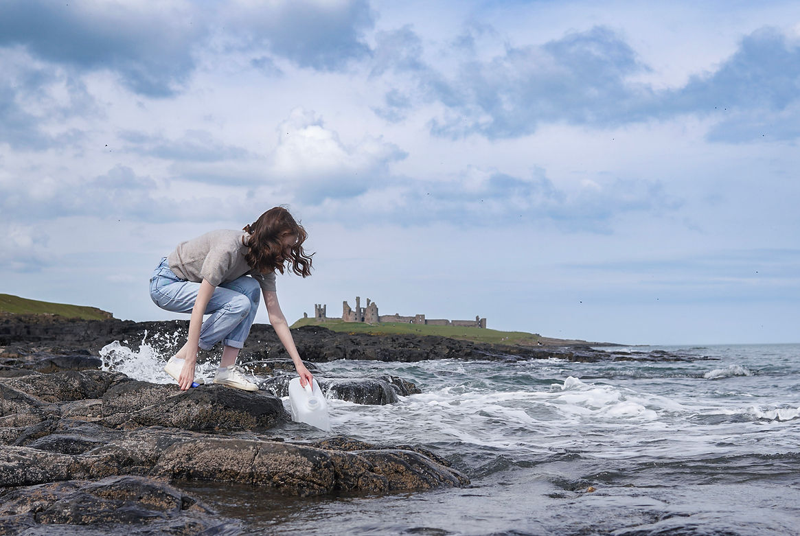 Woman collecting sea water on a rocky coastline with dunstanburgh castle in the background.