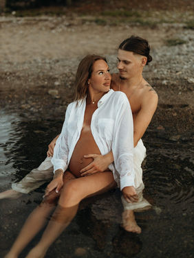 Gravidfotografering med ett par sittandes i vattnet och tittar på varandra iklädd ljusa kläder
