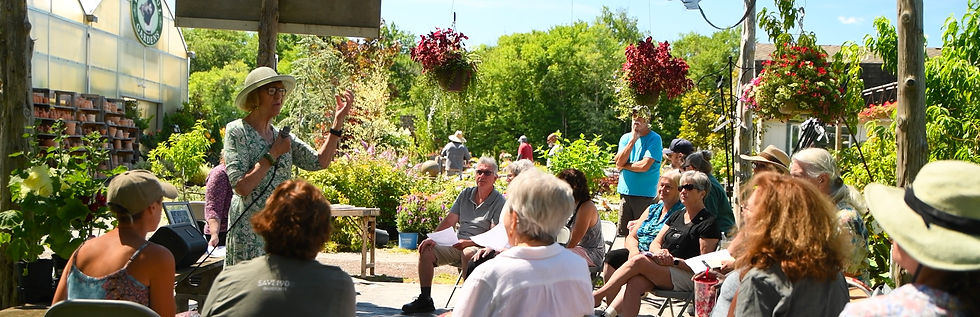 Woman in sunhat speaks to a seated group in a lush flower garden, with hanging baskets and bright greenery on a sunny day.