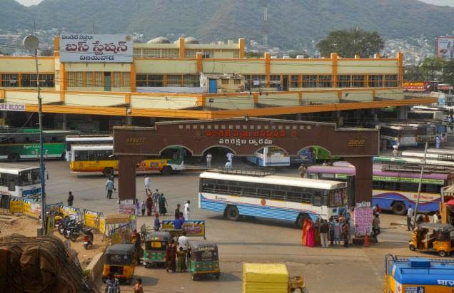 Busy bus station with colorful buses and autorickshaws. People gather near the entrance. Building with Telugu signage in the background.