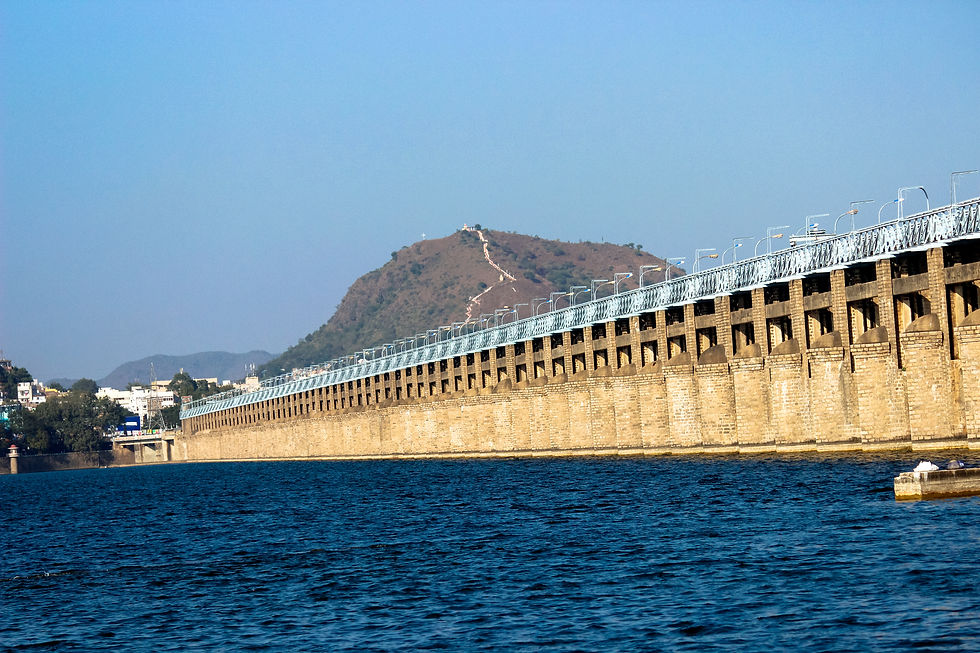 Large stone dam stretches across a river under blue sky. A hill with a winding path is visible in the background, creating a serene mood.