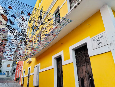 Edificio amarillo con puertas de madera bajo techo de encaje colorido. Letrero dice "Callejón del Oro". Ambiente alegre y soleado.